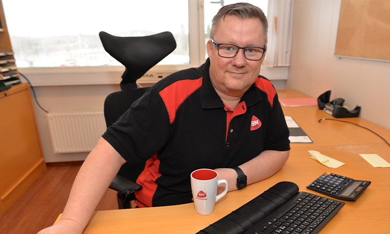 Ansatt i svart/rød BM-polo ved skrivebord med BM-kopp, PC-tastatur og kalkulator