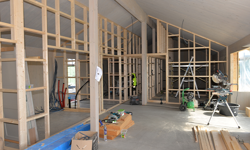 Interiør under oppføring med lyse trebjelker, stillas, sirkelsag og byggeverktøy.