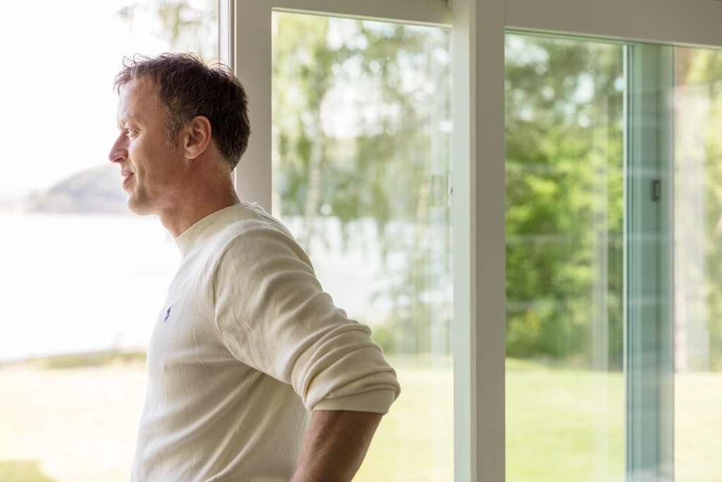 Mann i kremhvit genser i lyst rom ser ut av store vinduer mot grønt landskap og innsjø.