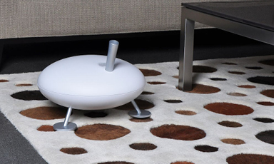 Hvit rund enhet med tre metallføtter og stang, på hvitt teppe med brune flekker ved sofa.