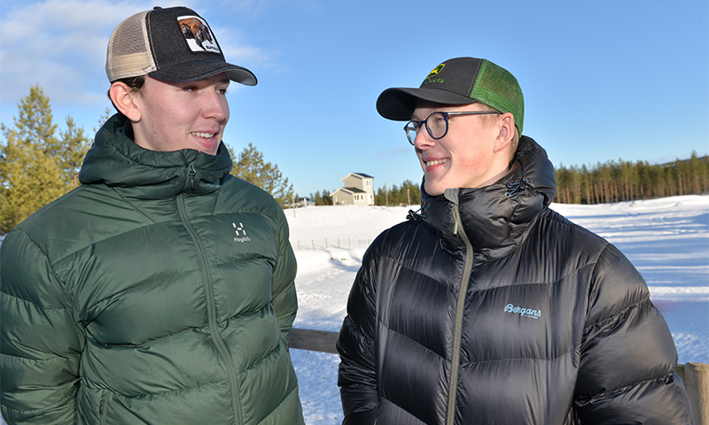 To menn i grønne og svarte dunjakker og caps ved snødekt gjerde foran hus og skog.