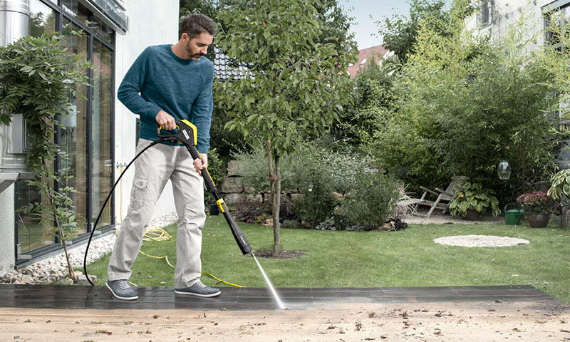 Mann vasker mørkt treborddekk med gul-svart høytrykksvasker i grønn hage.