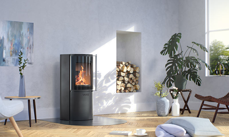 Moderne stue med svart rund vedovn i flammer, stabler med ved, lyse vegger og grønne planter.