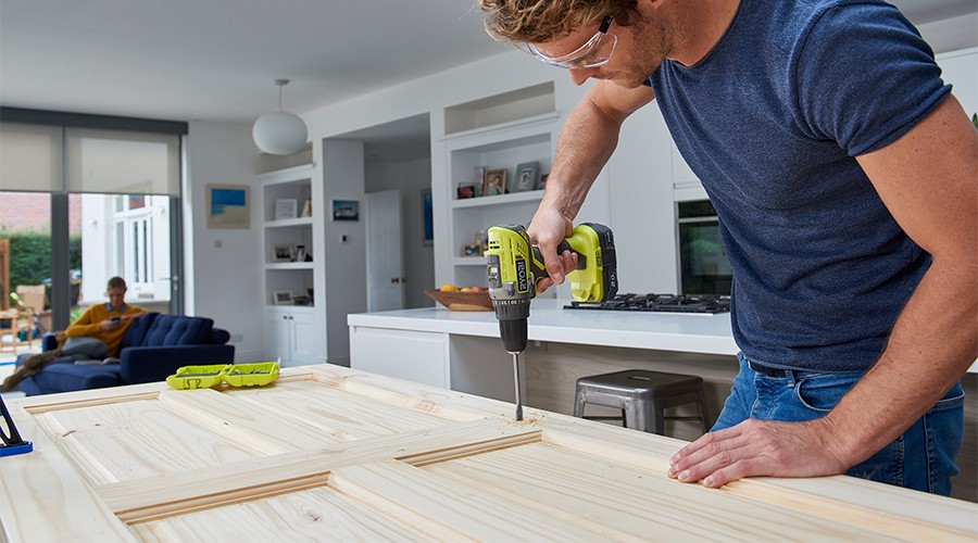 Mann med gulgrønn drill fester ubehandlet dør i hvitt kjøkken, bak person i blå sofa.