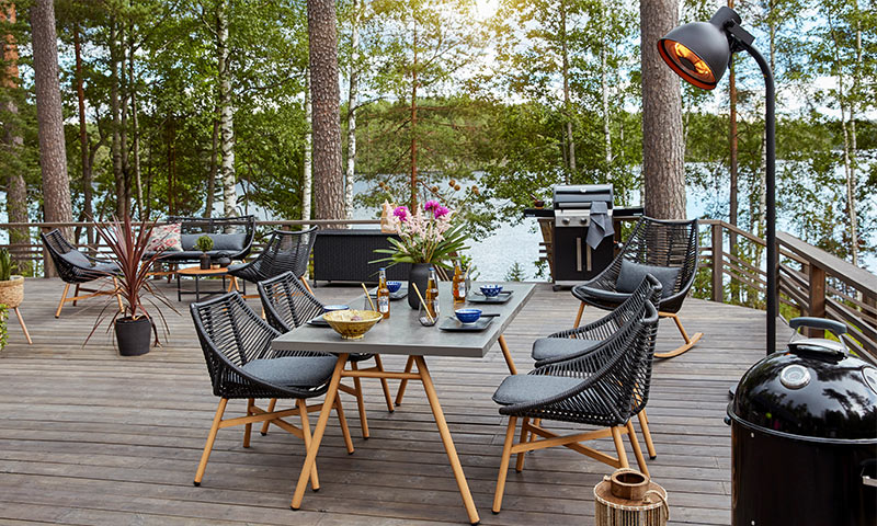 Møblert treterrasse med sort flettmøbler, grått bord, blomster, grill og innsjø- og skogutsikt