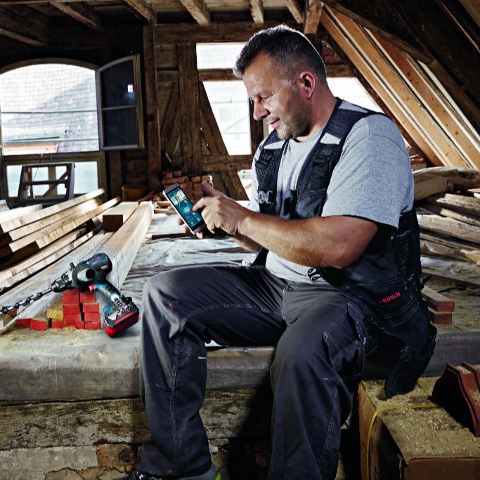 Håndverker i grå t-skjorte og svart vest på loft med treplanker, mobil og blå-rød drill.