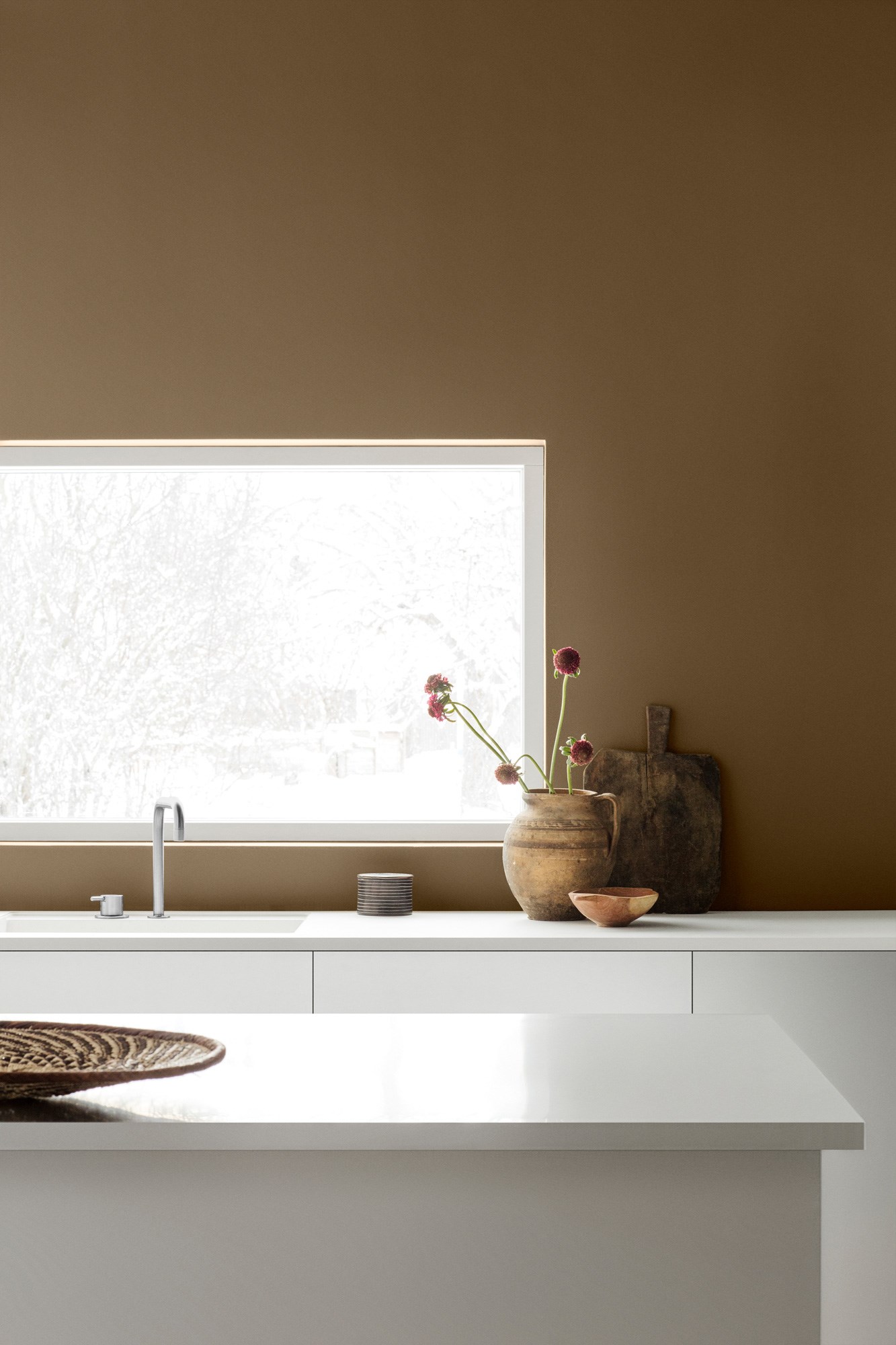 Minimalistisk kjøkken med hvit benk, brunt vegg, snøutsikt, leirkrukke med rosa blomster.