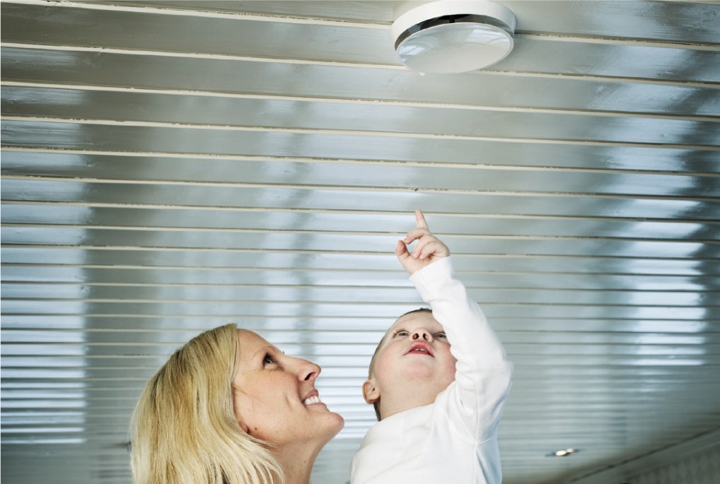 Voksen holder barn som peker mot hvit røykvarsler i sølvfarget tak