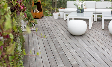 Lysegrå treterrasse med hvit loungegruppe, runde puffer og grønne potteplanter.