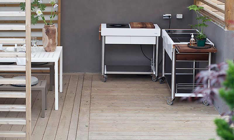 Lyst uterom med hvitt bord, vase med hvite blomster, og to mobile kjøkkenvogner foran grå vegg.