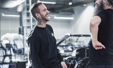 To menn i svarte klær ved en motorsykkel i et lyst, industrielt verkstedmiljø.