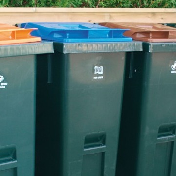 Tre grønne avfallsbeholdere med blå, oransje og brunt lokk foran et tregjerde