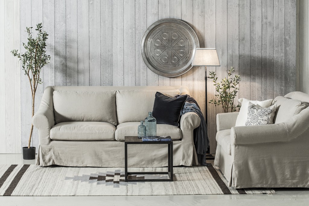 Lysegrå sofa og lenestol med blå puter, hvit plankevegg, rund metalldekor, lampe og planter