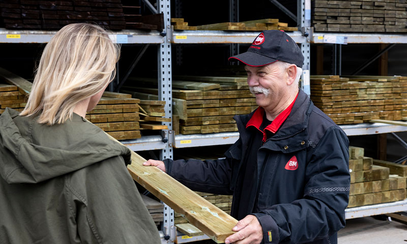 Ansatt i svart jakke gir trykkimpregnert treplanke til kvinne i grønn jakke foran hyller.
