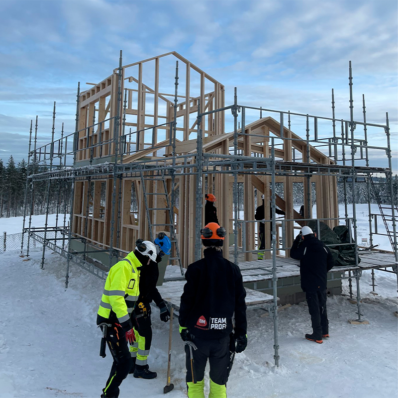 Trekonstruksjon omringet av grå stillas, arbeidere i gule og oransje jakker i snødekt skog