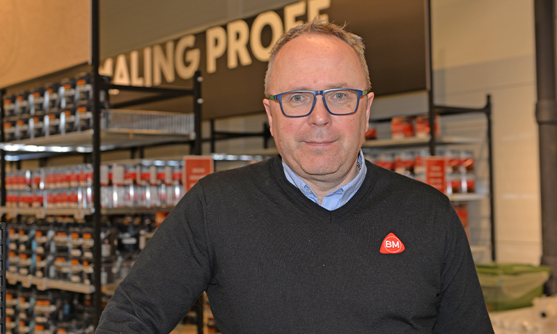 Ansatt i blå skjorte og svart genser med rødt BM-merke foran hylle med malingsspann