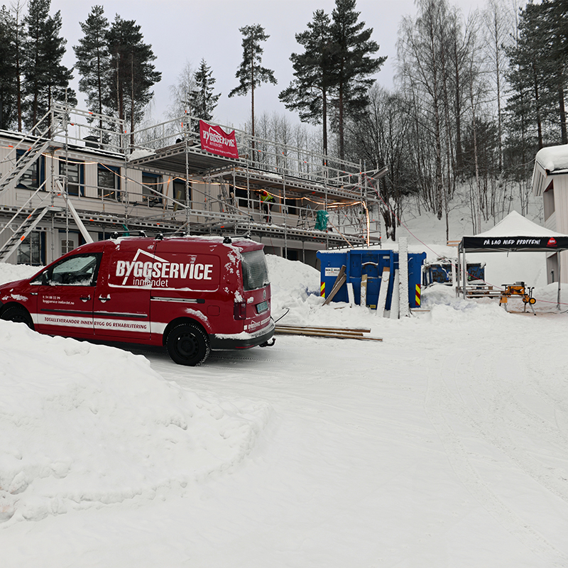 Rødt Byggservice-kjøretøy foran hvit, stillasbygget bolig i snødekt skoglandskap.