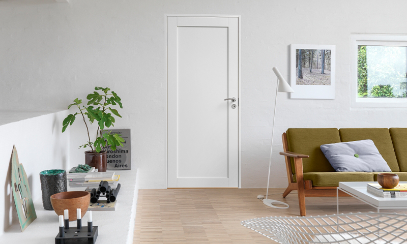 Minimalistisk stue med hvit dør, lyse tregulv, grønn sofa, plante og veggkunst.