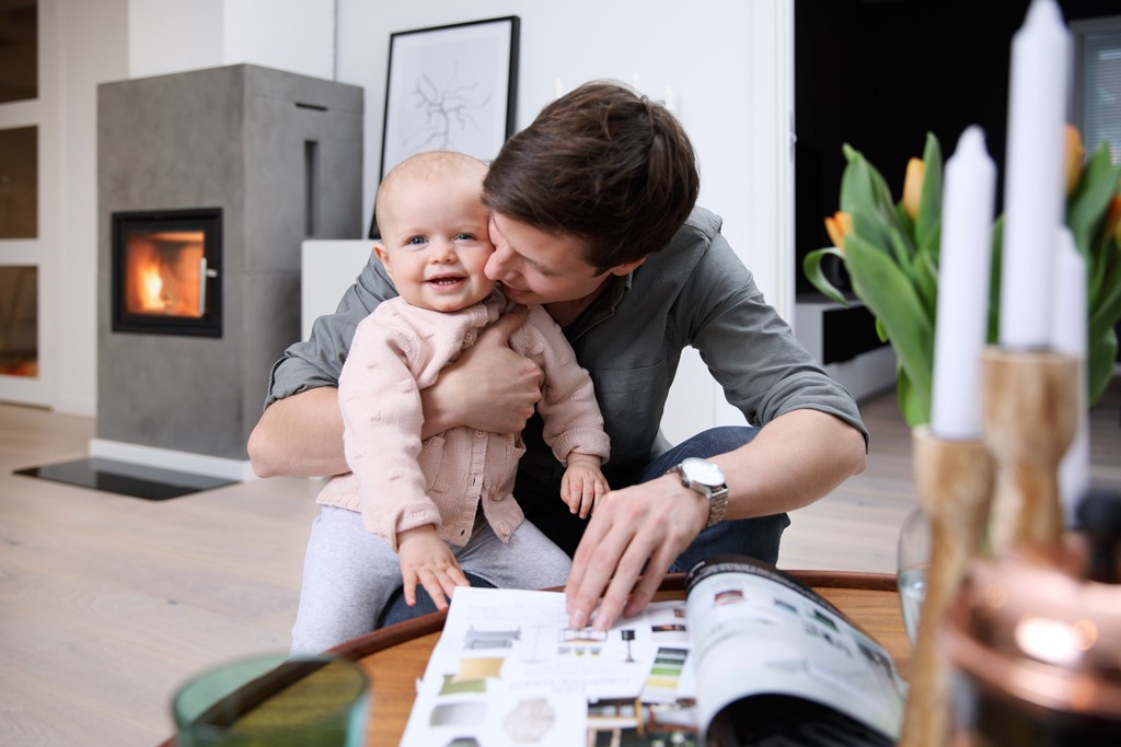 Far holder barn på fanget og blar i interiørkatalog ved levende peis i lys stue med tulipaner
