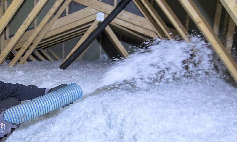 Blåstripet slange blåser hvit løsfyllsisolasjon i loft med synlige trebjelker.