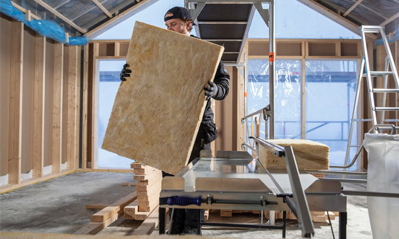 Arbeider i råbygg bærer gul isolasjonsplate foran sagbord og trestendere.