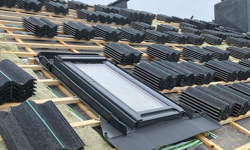 Skråtak under bygging med stabler av svarte takstein på lekter og nytt takvindu.