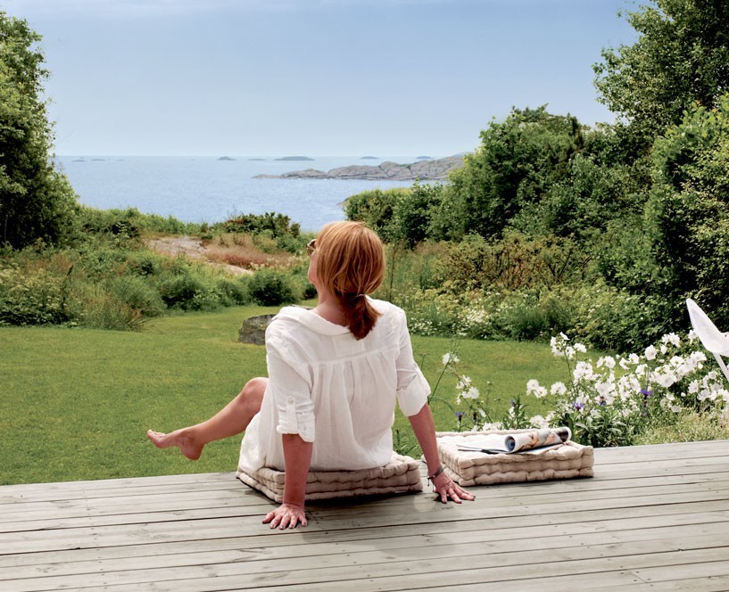 Rødhåret kvinne i hvit bluse sitter på tredekk med sjøutsikt, grønn hage og hvite blomster.