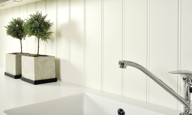 Hvit kjøkkenbenk med synlig vask og kromkran, hvit panelvegg og to små prydtrær i grå betongpotter.