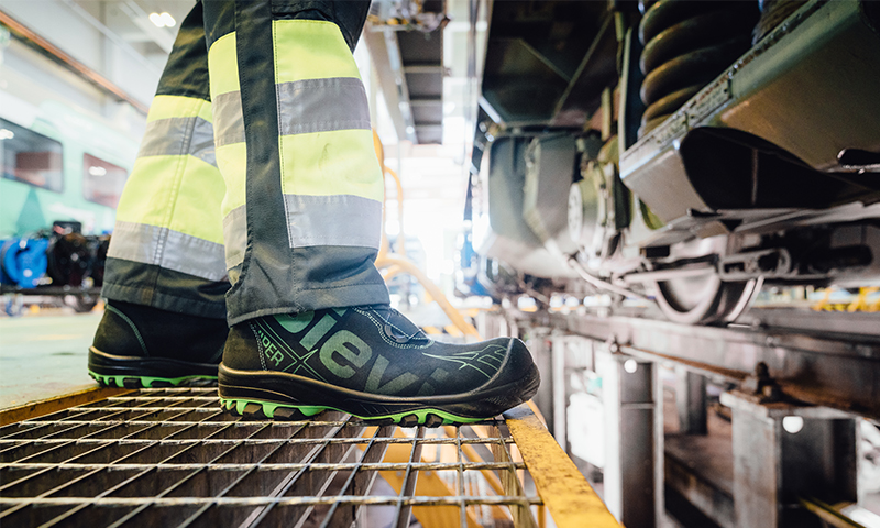 Nærbilde av svart-grønn vernesko og neongule refleksbukser på gittergulv ved togspor.