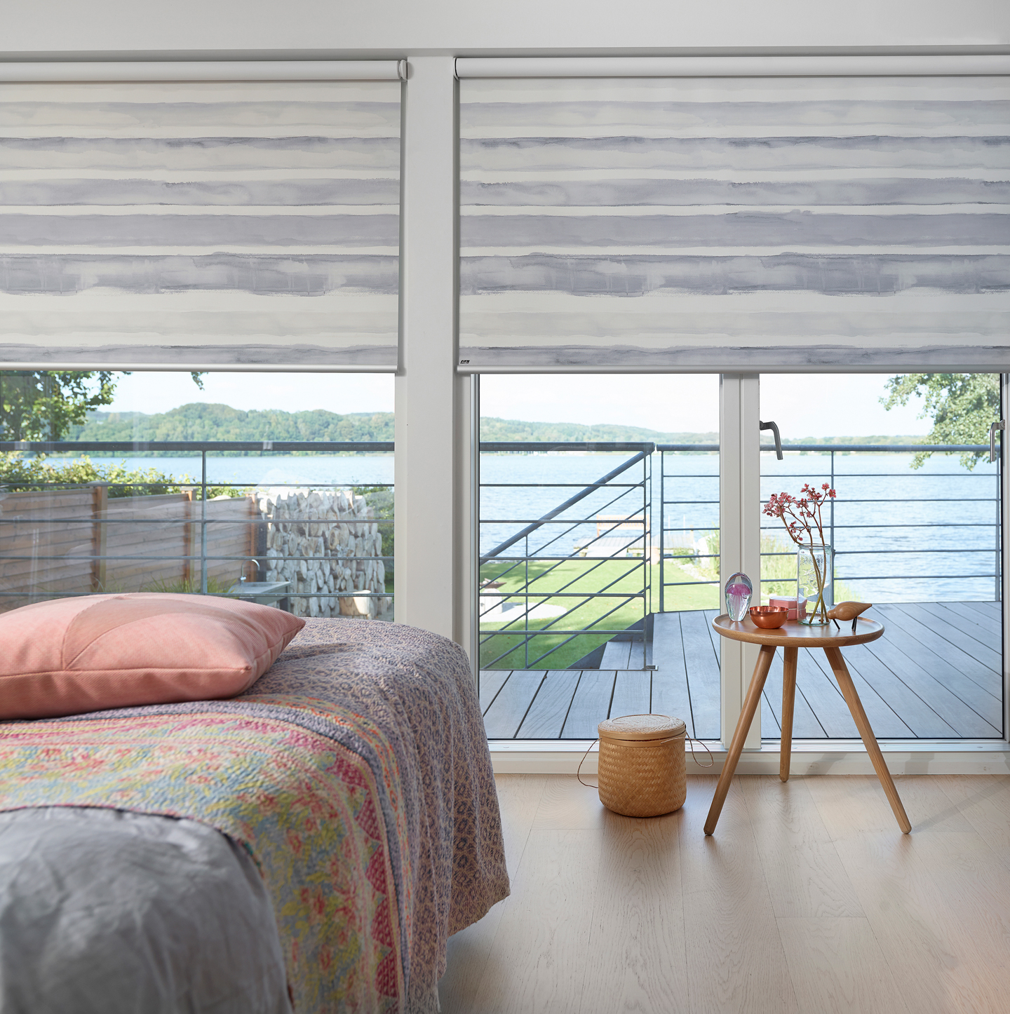 Lyst soverom med mønstrete sengeteppe, rosa pute, gråstripete rullegardiner og innsjøutsikt.