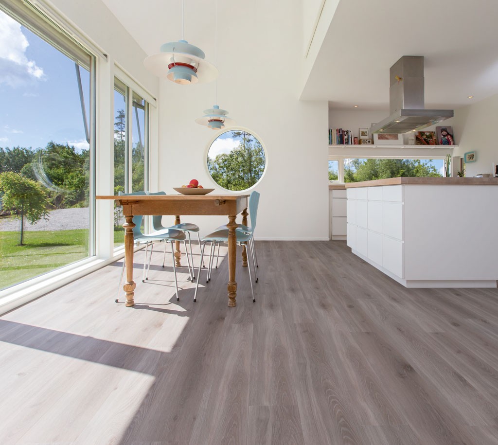 Lyst, moderne kjøkken med store vinduer mot grønt ute, rund luke, trebord, lyseblå stoler.