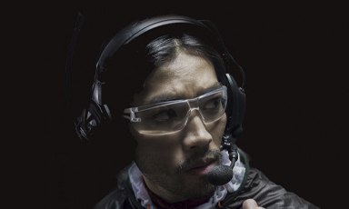 Person med mørk hettegenser og store svarte hodetelefoner mot svart bakgrunn.