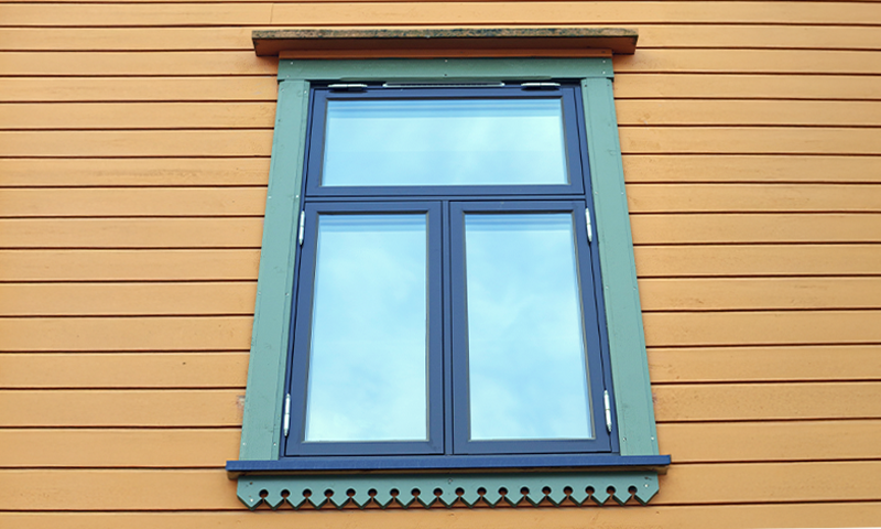 Vindu med blå ramme og dekorativ grønn list på gul trefasade, reflekterer blå himmel.