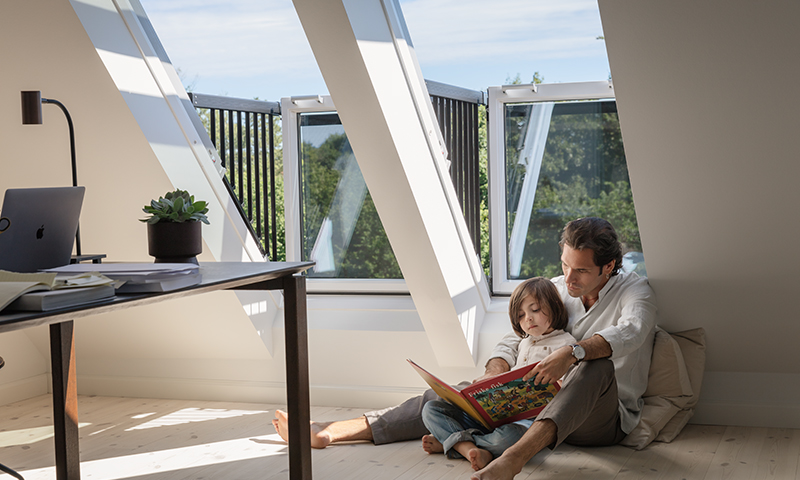 Forelder og barn leser bok på tregulv ved åpne takvinduer i lyst loftsrom.