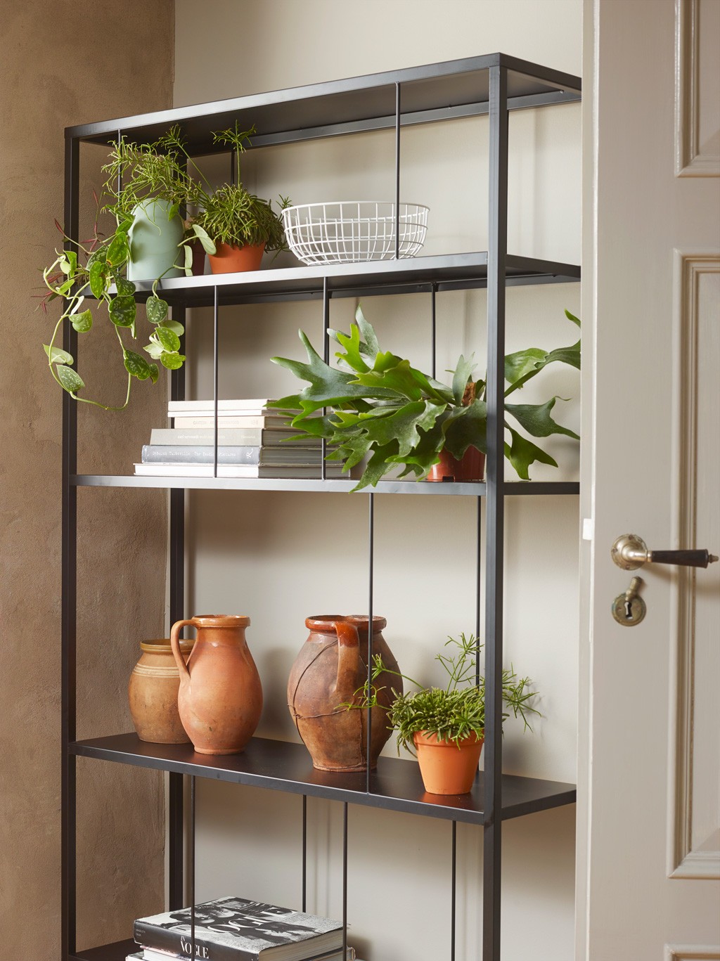 Sort metallhylle mot beige vegg med terrakotta, grønne planter, bøker og hvit trådkurv.