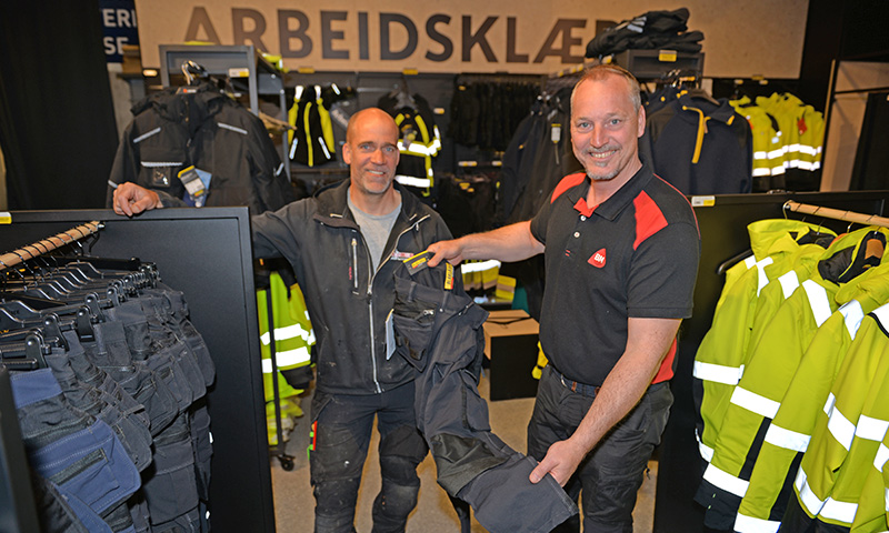 To menn i arbeidsklærbutikk under skilt ARBEIDSKLÆR, holder mørke bukser, gule refleksjakker bak