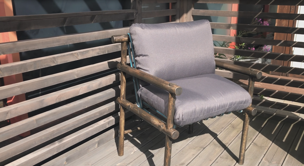 Rustikk stol i tre med grå pute på solfylt terrasse foran trepanel.