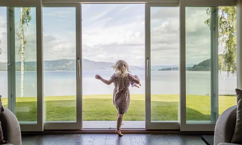 Lite barn i blomstrete kjole med dukke går gjennom glassdører mot grønn plen og blå innsjø