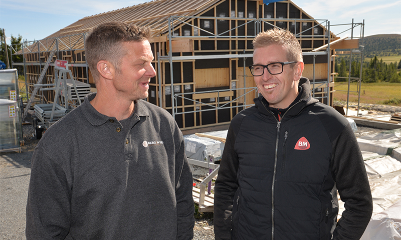 To menn ved hus i reisverk med stillas, byggevarer, grønne åser og blå himmel.