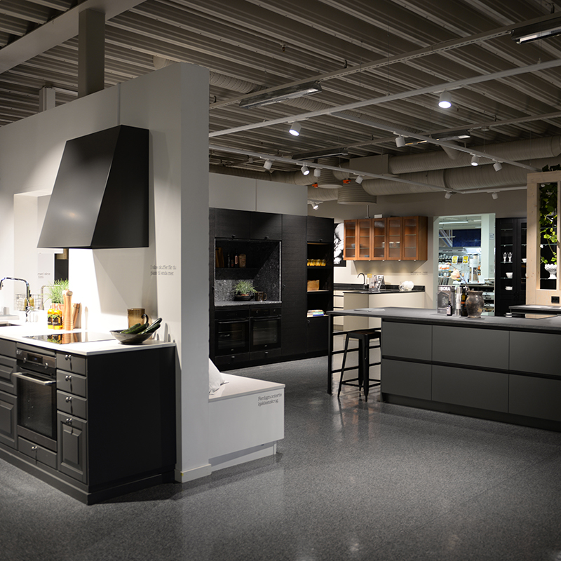 Moderne kjøkkenutstilling i butikk med svarte og grå skap, hvit benkeplate og trehyller.