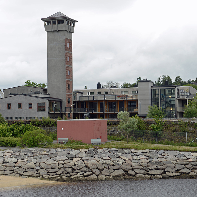 Grå bygning med høyt tårn ved steinlagt vannkant, to benker og rødt skjul i forgrunnen.