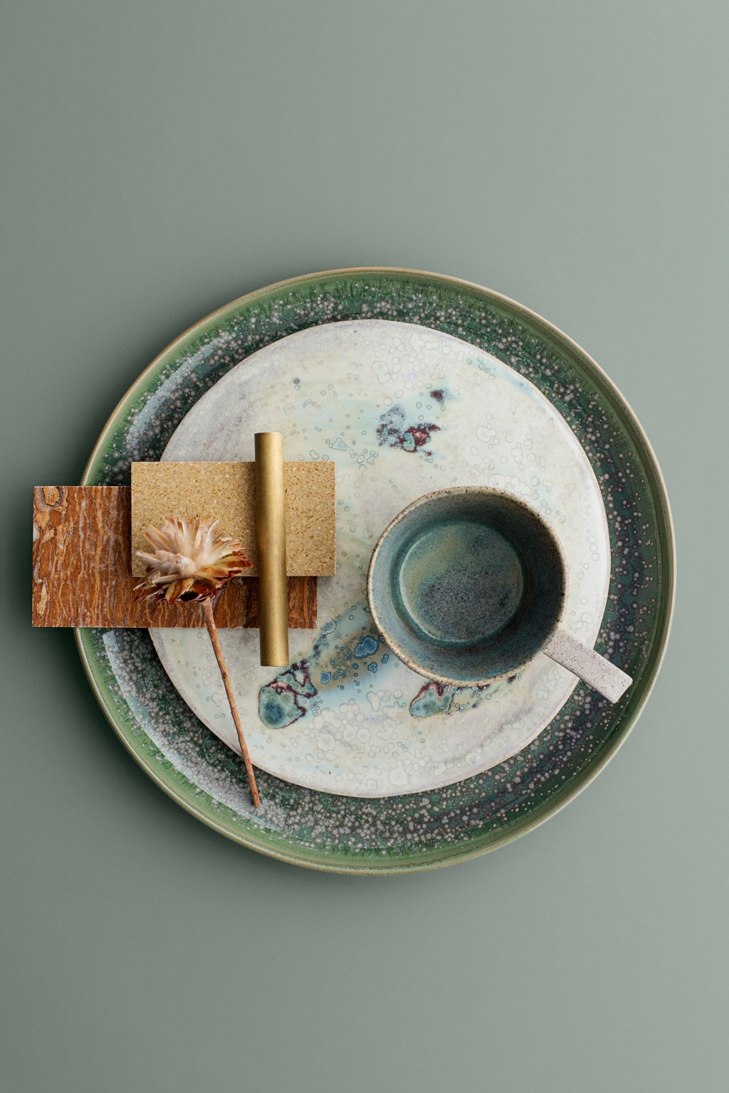 Flat oppstilling med grønt spraglete fat, kopp, gullstang, treplater og tørket blomst