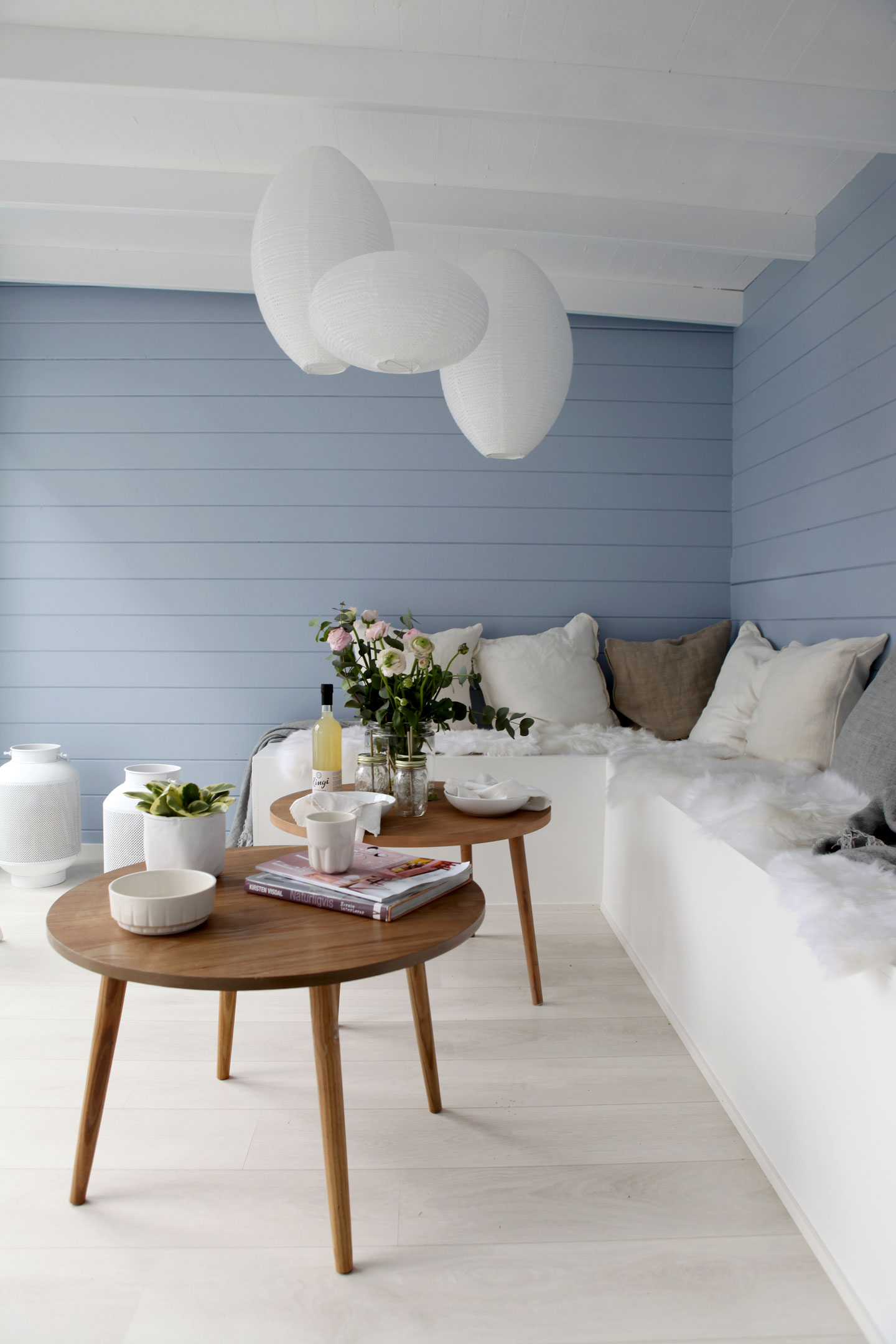 Koselig hjørne med lyseblå panelvegg, hvite papirlanterner, runde trebord og puter.