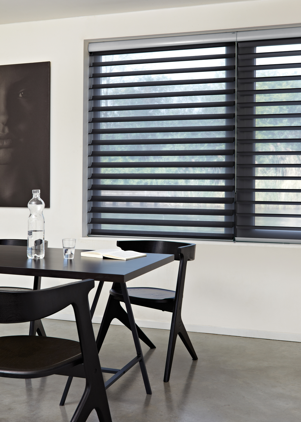 Svart spisebord med to stoler ved vindu med mørke persienner, vannflaske, glass og åpen bok.