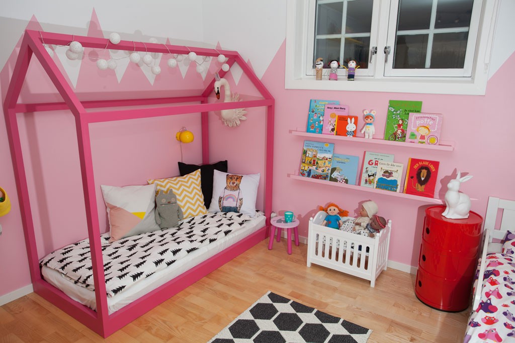 Rosa barnerom med husrammeseng, bokhylle med fargerike barnebøker og rødt skuffeskap.