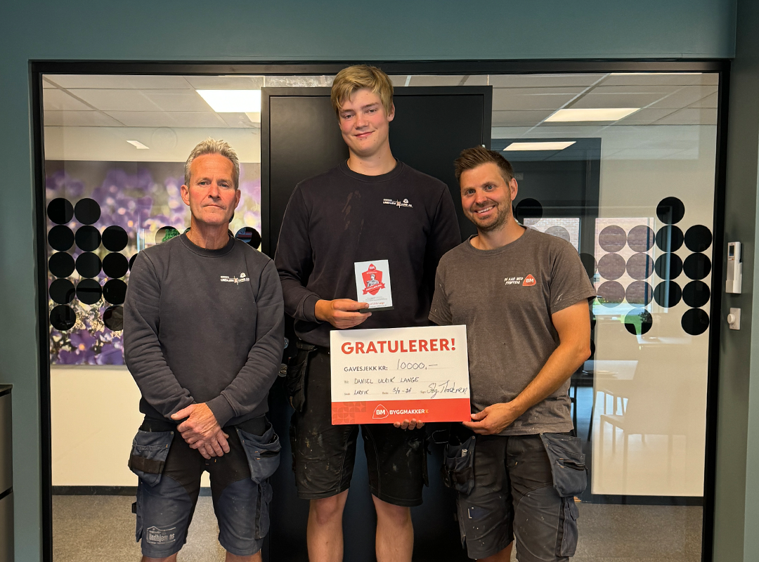 Tre håndverkere i mørkt arbeidstøy holder hvit-rød sjekk med Gratulerer!-tekst inne i butikk.