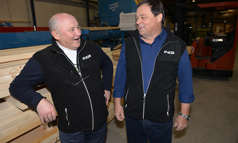 To menn i svarte vester foran stabler med lys trelast og rød truck i industrilokale.