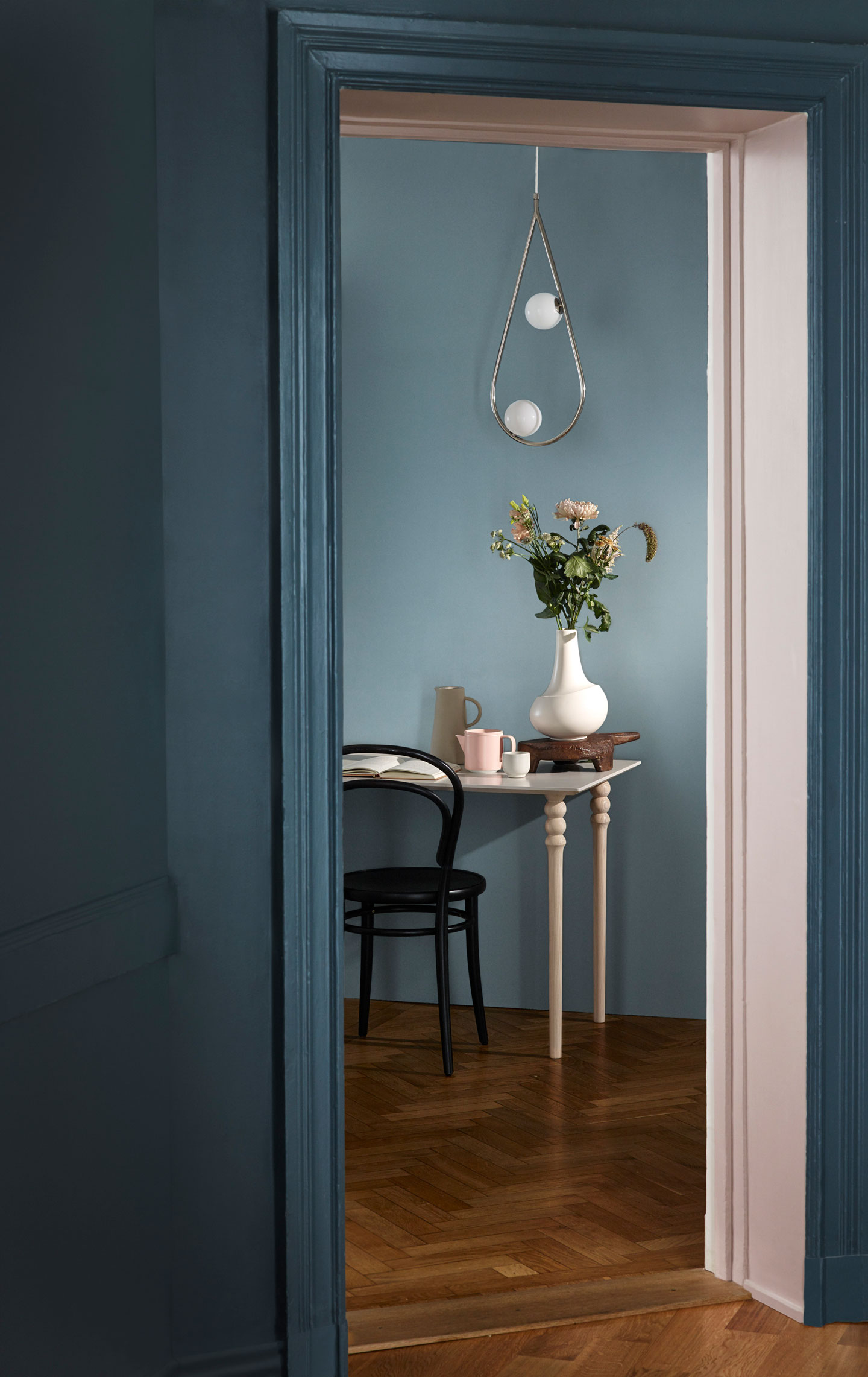 Dørblikk inn til blå rom, hvitt bord med vase og blomster, rosa kopp, hengende dråpelampe.