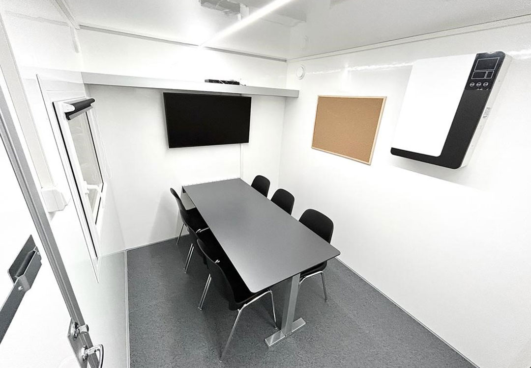 Lite hvitt møterom med grått bord, seks svarte stoler, vegg-TV, korktavle og luftrenser.