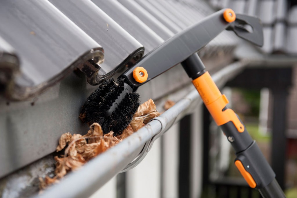 Grå-oransje teleskopstang med svart børste fjerner brune blader fra grå takrenne.